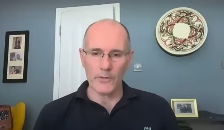 Professional speaking during a video call from a home office setting, facing the camera during an online meeting or presentation.
