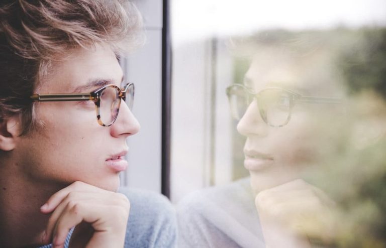 Person wearing glasses looking thoughtfully out of a window, with their reflection visible in the glass.