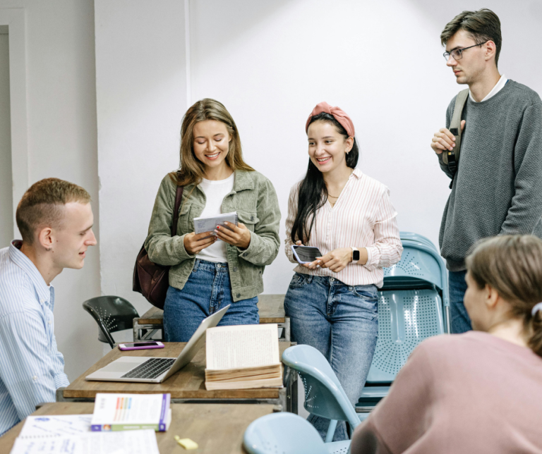 Group of young adults collaborating in a classroom or workshop setting, smiling and engaging in discussion around a desk with a laptop and study materials.