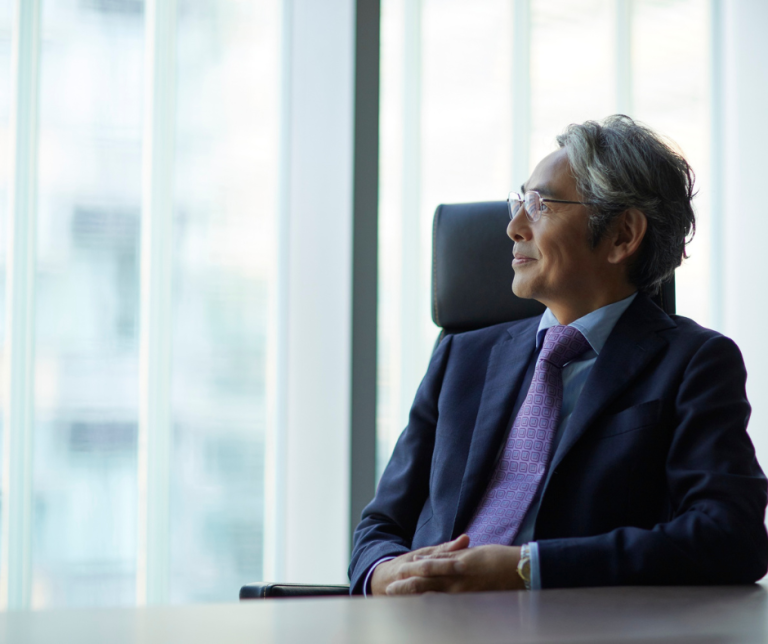 Professional in business attire seated in an office, looking out of a window in a thoughtful or reflective moment.