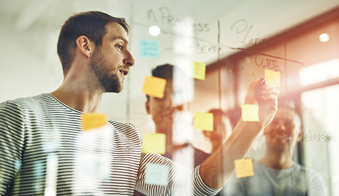 Professional leading a brainstorming session, placing sticky notes on a glass board while collaborating with colleagues in an office setting.